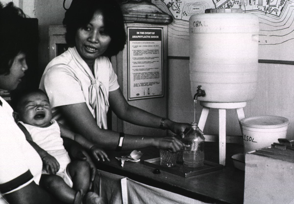 A woman shows water level in a bottle to fill as another woman looks on holding a crying baby.
