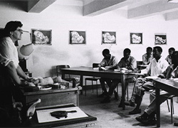 A woman instructor stands facing several men with books seated by desks in a classroom. 