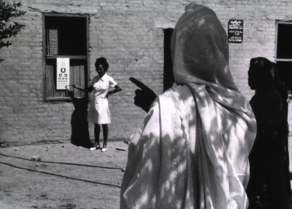 A woman stands and points to an eye chart while a person stands in the foreground taking the test. 