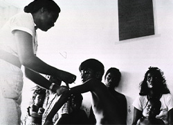 A nurse stands and gives an injection to a seated boy’s upper left arm as others look seated in the background.