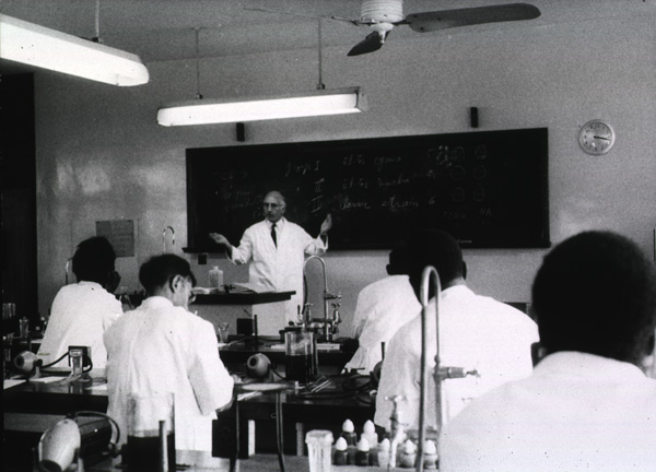 In a laboratory classroom, a man lectures to several who are seated. 