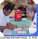 Man in white medical coat holding surgical instrument sitting across from student holding medical instruments.  Courtesy Kenneth Davis, Jr., M.D.