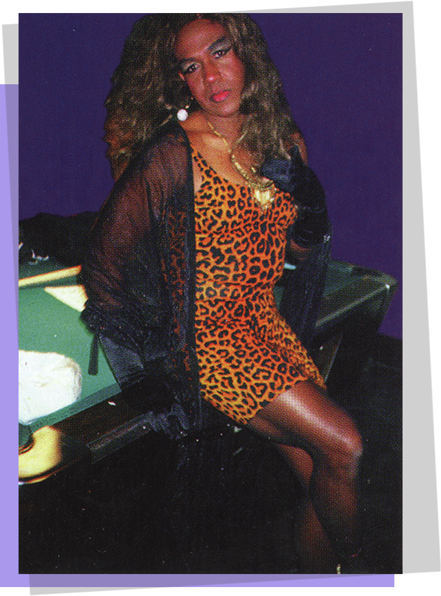 An African American male wearing a leopard print dress and black shawl while leaning on a pool table
