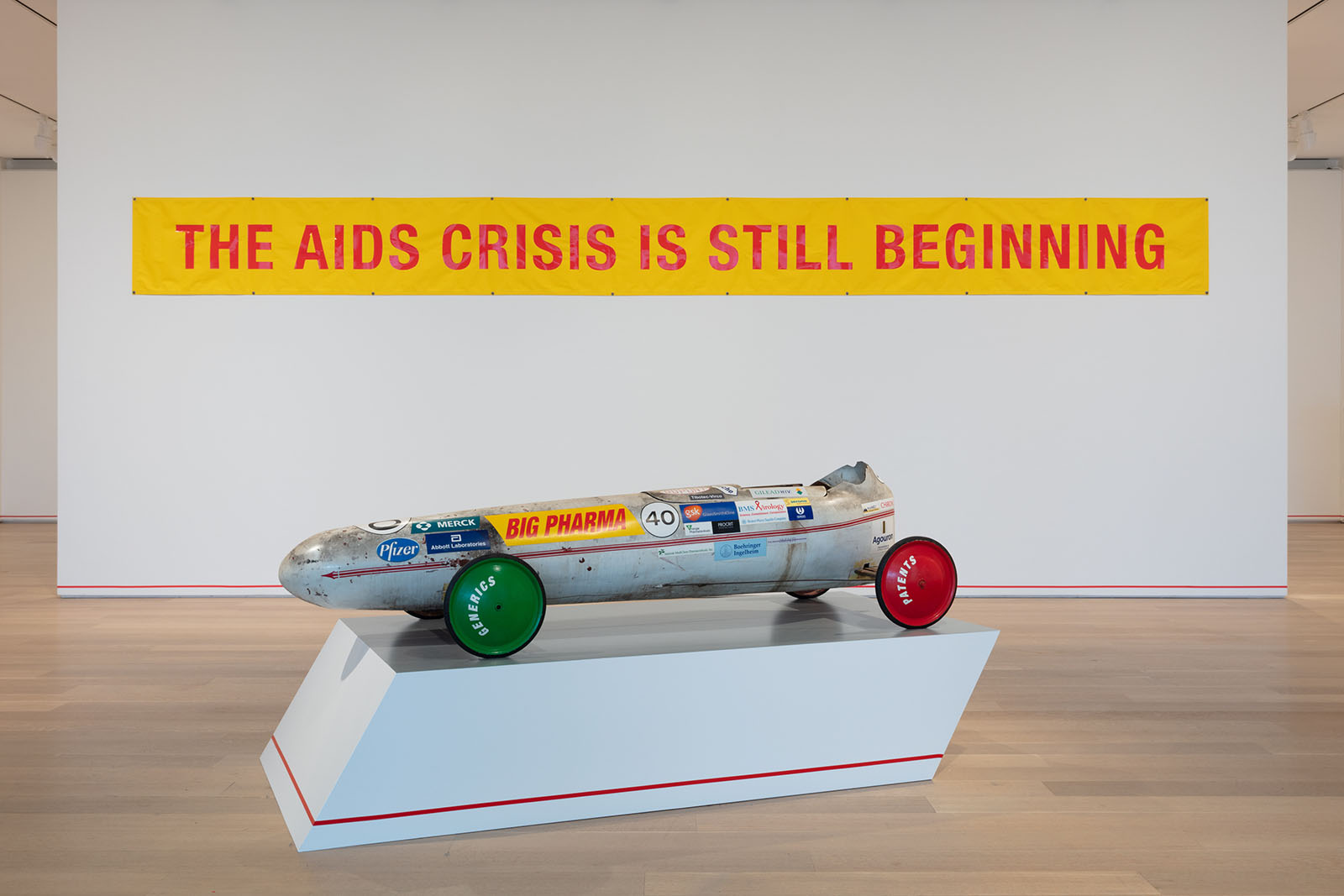 A gallery installation with a sculpture and on the back wall a yellow poster with red text that says “The AIDS Crisis is Still Beginning”