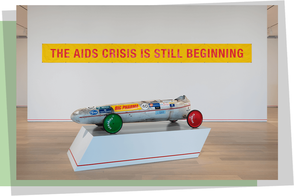 A gallery installation with a sculpture and on the back wall a yellow poster with red text that says “The AIDS Crisis is Still Beginning”