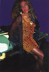 Image of an African-American male dressed in a leopard skin dress and wearing make-up leaning on the hood of a green car.