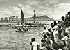Black and white photograph, the Hōkūle‘a voyaging canoe coming into harbor after its inaugural voyage as spectators watch. 