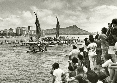 Black and white photograph, the Hōkūle‘a voyaging canoe coming into harbor after its inaugural voyage as spectators watch.