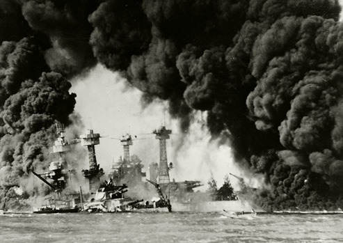 In this black and white photograph, battleships are surrounded by black smoke during the attack on Pearl Harbor.