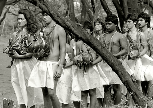 A procession of people participating in the Makahiki ceremony stand amongst trees. Each person wears a white skirt and many carry ceremonial objects.