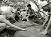 7 people stand around an adze grinding stone on Kaho‘olawe. A woman and man in the foreground touch the stone, white the rest stand a small distance from it. Trees are in the background. This is a black and white photo.