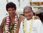 Nainoa Thompson (left) of the Polynesian Voyaging Society stands next to Mau Piailug (right), Micronesian wayfinder in front of boat on beach.