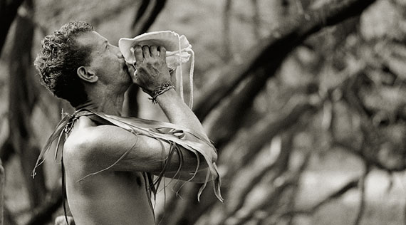 Black and white photograph of man with palm fronds around his neck blows into a conch shell during the Makahiki.
