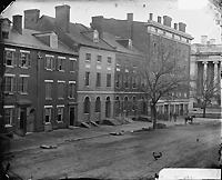Black and white photograph of several brick buildings in a row with a few people and a horse in the background. Courtesy Library of Congress.