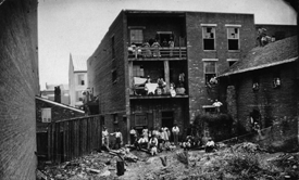 Black and white photograph of a brick building showing African American women on two balconies with hanging laundry.  African American women and men along with white men and children can be seen sitting and standing in the courtyard below. Courtesy National Archives, Washington, D.C. 