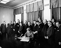 Several white men and one woman standing behind another man at a desk signing a paper.