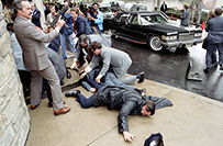 Two men injured lie on the ground while others surround them. One man standing in front holds a gun and looks on to the right.