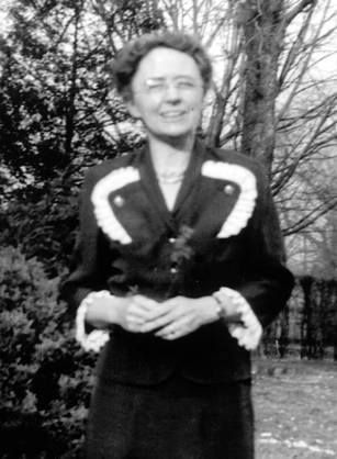 Dr. Emily Partridge Bacon, a White woman in a blouse and skirt standing outdoors near shrubs.