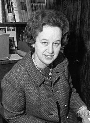 Dr. Marianne Schuelein, a White woman posing at a desk with lots of papers and a bookshelf in the background.