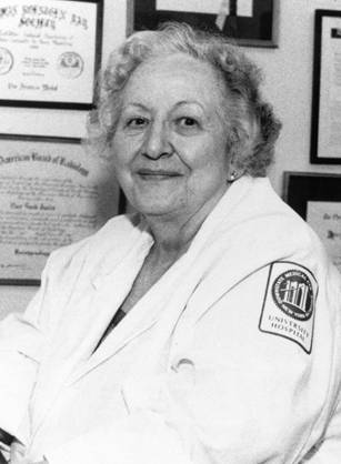 Dr. Lucy Frank Squire, a White woman seated at desk in a lab coat with medical degrees hanging on the walls.