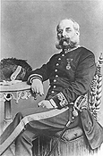 Full length, black and white photograph of S. L. Pisani seated in a chair wearing a military uniform of the Chief Government Medical Officer of Malta with a mustache and a beard. His left leg is crossed over his right leg. His right arm is resting on a table.