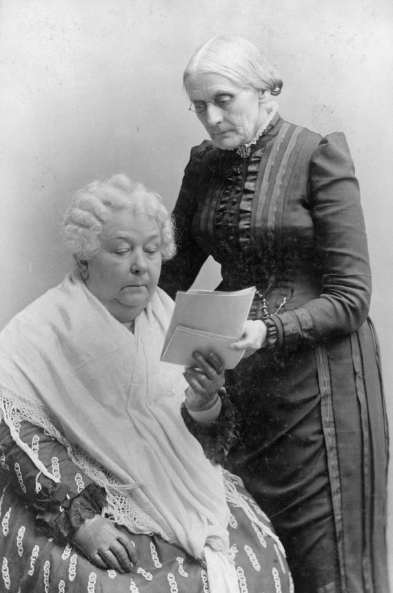 An old White woman sits and reads a pamphlet, another old White woman stands over her left shoulder.
