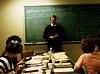 A White man stands in front of chalkboard facing 6 women seated around paper filled table.