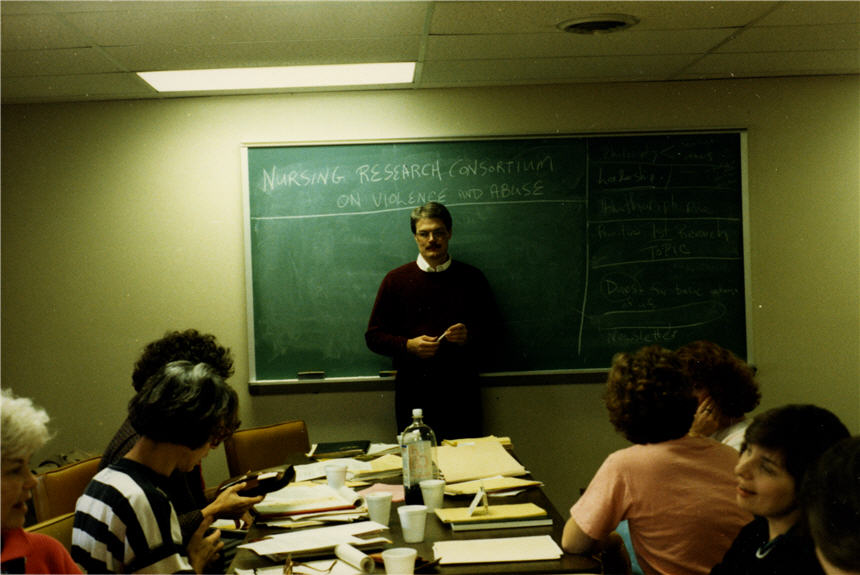 A White man stands in front of chalkboard facing 6 women seated around paper filled table.