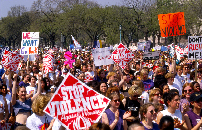 A large crowd stands on the Mall facing right, many hold signs.