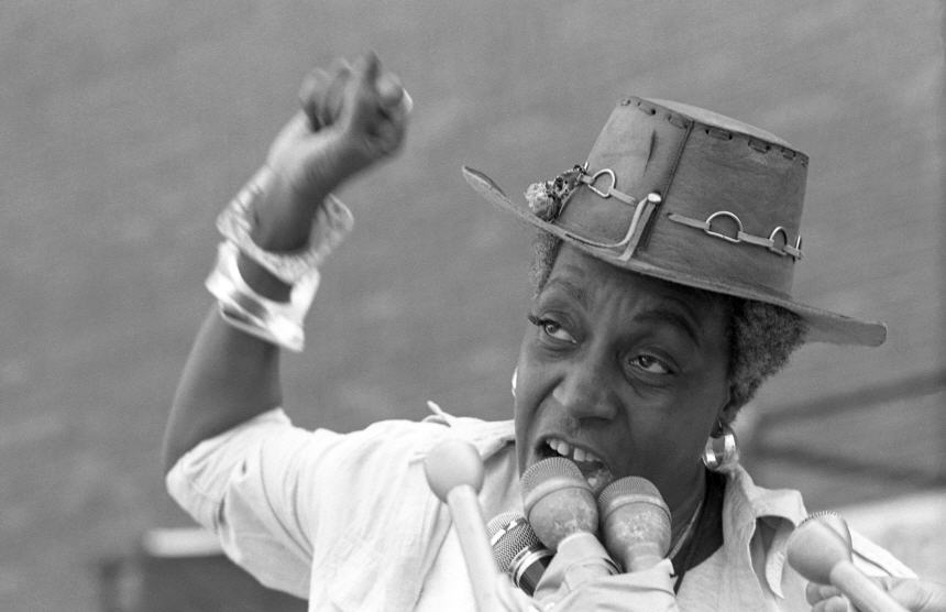 African American woman speaks in front of a bank of microphones to a crowd out of view to the left.