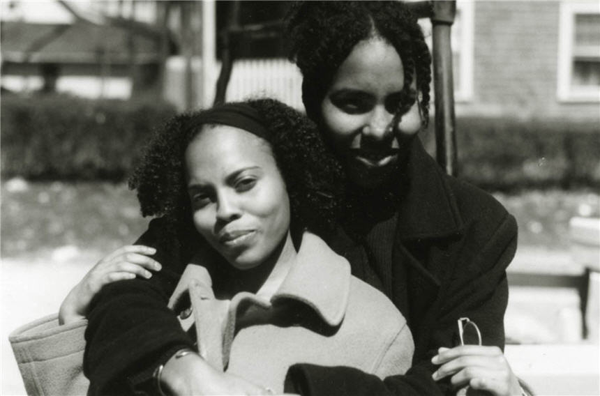 Two African American women seated and facing viewer, woman on left front leans into woman behind.