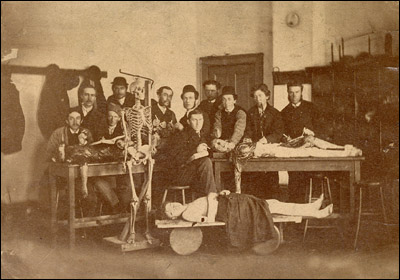 Cropped photograph of the interior of an unidentified dissecting room, students pose next to three cadavers and a skeleton. United States, ca. 1910.