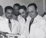 Black-and-white photograph of Charles Drew standing by a patient's bed and teaching twenty or so interns and residents during rounds at Freedmen’s Hospital, ca. 1947.