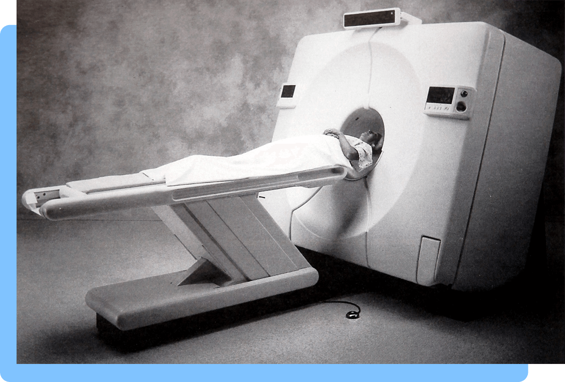 A patient lying on a flat examination table in front of the center of a large medical imaging device resembling a doughnut-like shaped machine