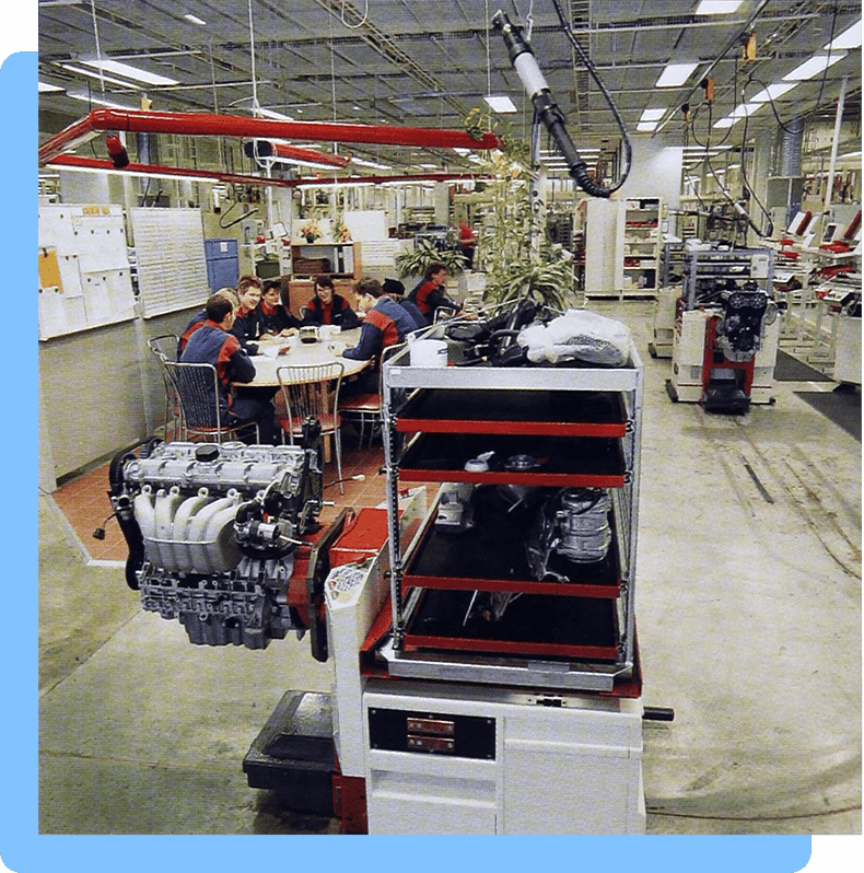 A group of workers sit at a table in a factory surrounded by automobile parts, equipment, and machinery