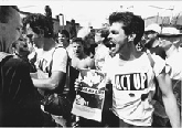 A black and white post card featuring a group of men wearing Act Up shirts. The man in the forefront is holding a poster in his right hands with the words Read my Lips on it.