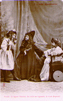 A black and white photograph post card with the title BeBe Apothicaire at the top and Voyez, la figure s'eclaire, les traites se reposent, la curse disaparait. The photograph is of three little girls playing doctor dress up. The girl on the left talks to a girl wearing a doctor's robe and hat while the third girl sits in a chair with wheels on it.