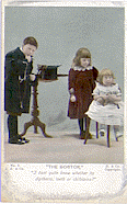 A hand colored post card with the title The Doctor, I don't quite know whether its diptheria, teeth, or chiblains. It features a boy wearing a dark robe holding his chin with his right hand and a stethoscope is draped over his arm. A girl stands behind another girl who is seated in a chair. 