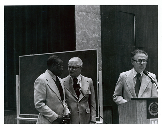 A black man talking with two white men next to a chalkboard and podium.