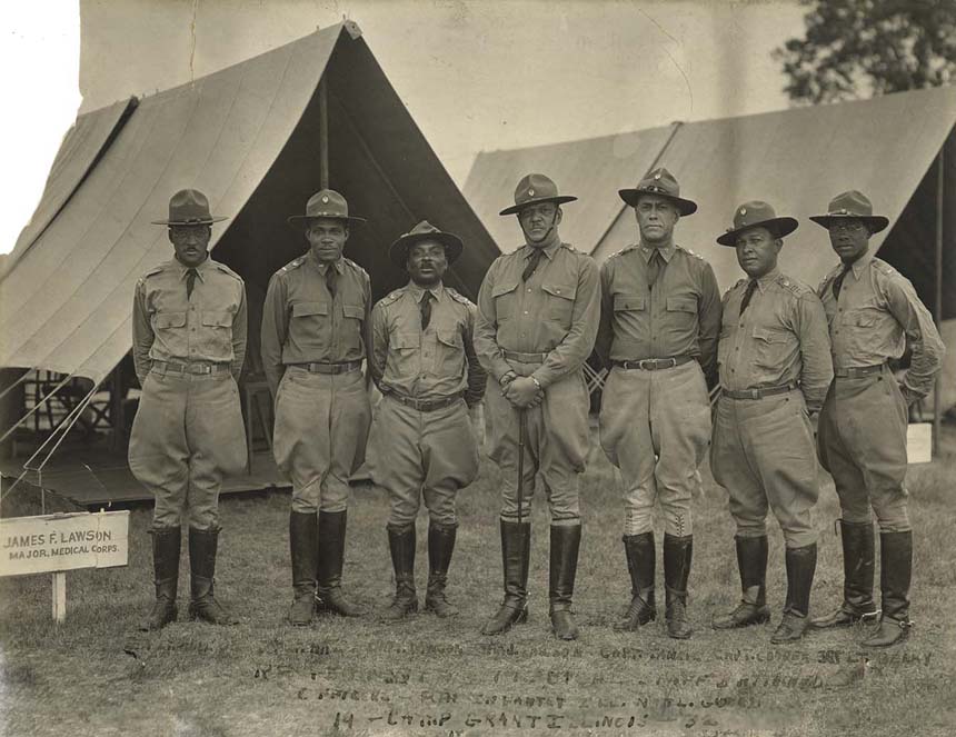 Photo of soldiers in uniform with hats and boots standing in a line in front of outdoor camp tents.