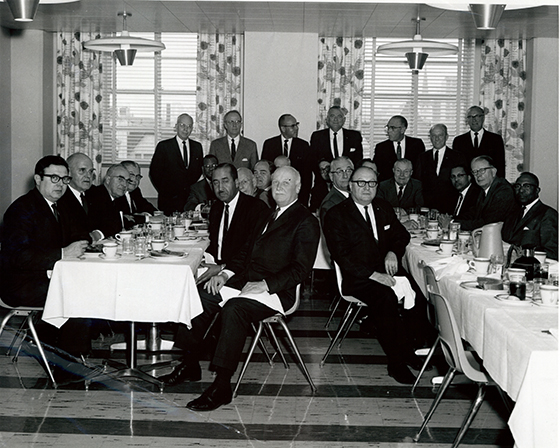 Diverse group of well dressed men sitting at two lunch tables.