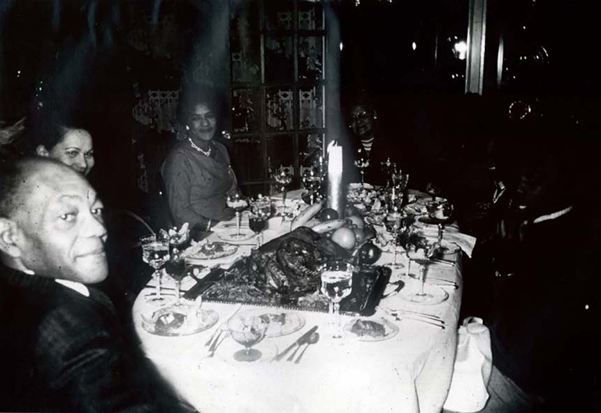 People seated at a table with a long tablecloth, wine glasses, a fruit centerpiece, and a cooked turkey.