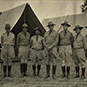 Photo of soldiers in uniform with hats and boots standing in a line in front of outdoor camp tents.