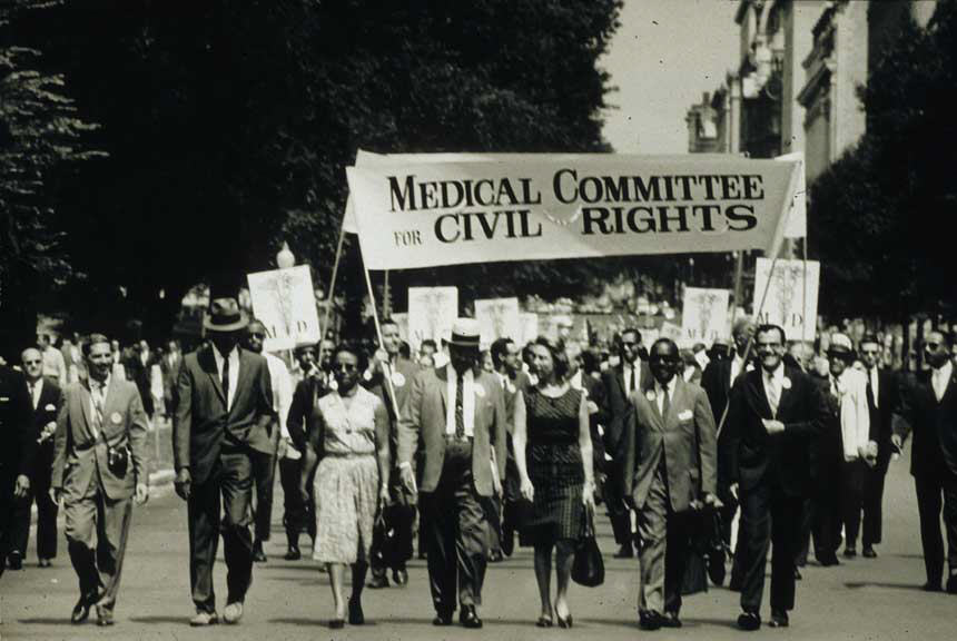 Large multiracial group of men and women holding signs and marching on a street towards viewer.