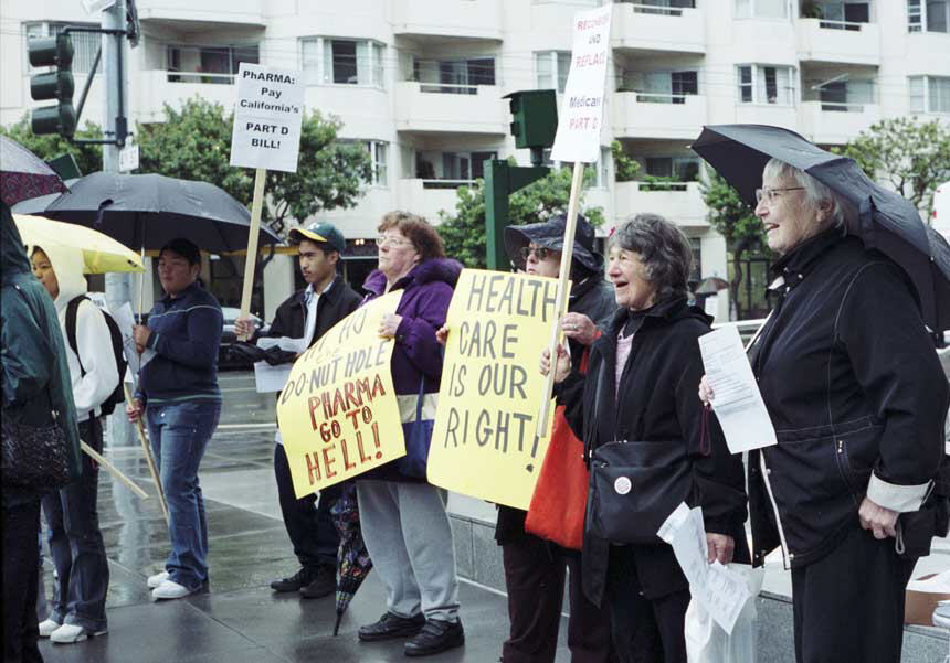Multiracial group of protesters in rain, holding signs, protesters in foreground are elderly.