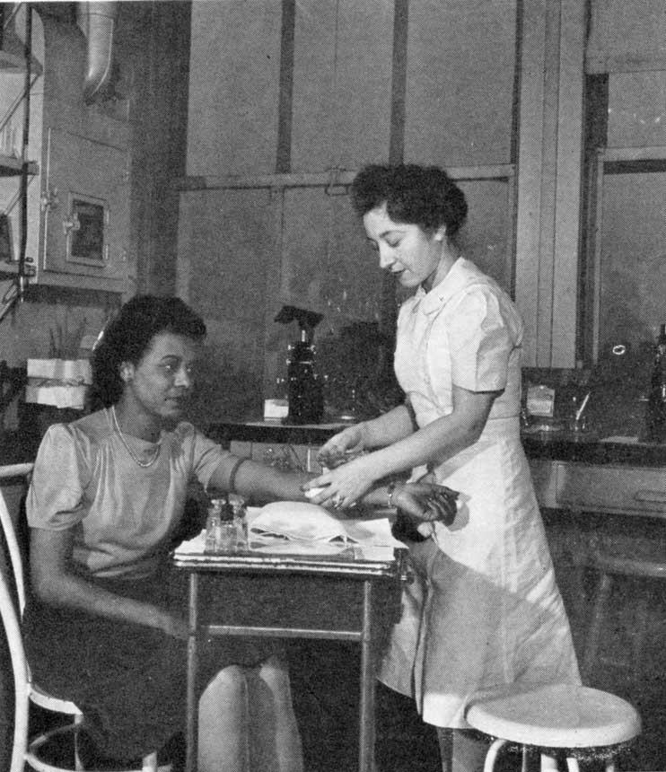 Standing White female nurse holds the arm of a seated African American woman.