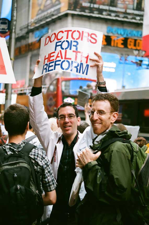 Two White male doctors smile facing viewer amidst a group of demonstrators in a street.