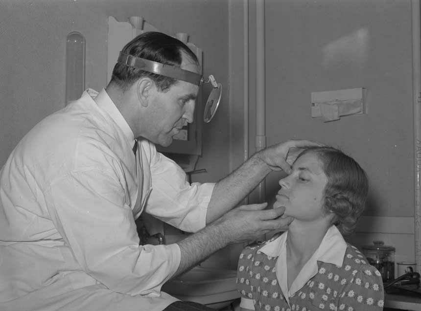 Standing White male doctor examines the face of a seated White woman.