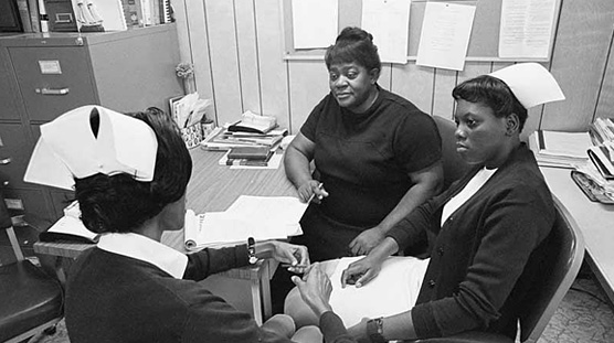 Three African American women sit around a desk; two of the women are nurses.