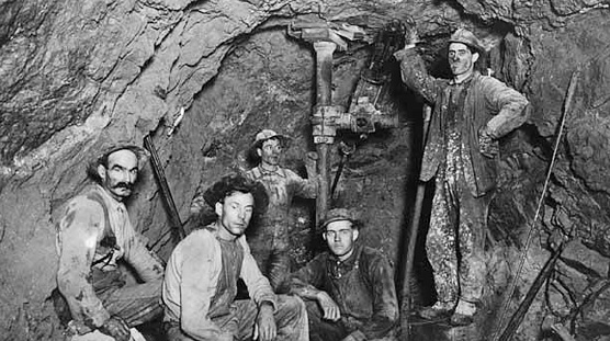 5 White male miners crouched and standing next to mining equipment underground.